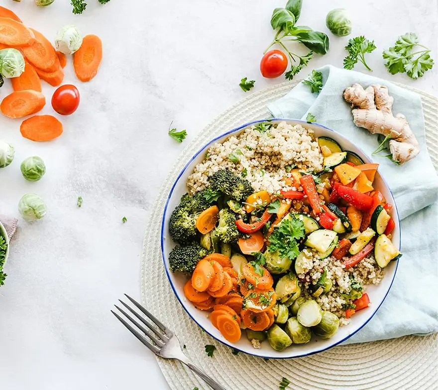 Colorful vegetable bowl featuring broccoli, carrots, zucchini, and cherry tomatoes, served with quinoa on a white plate. Fresh herbs and additional vegetables are scattered around, creating a vibrant and healthy meal presentation.