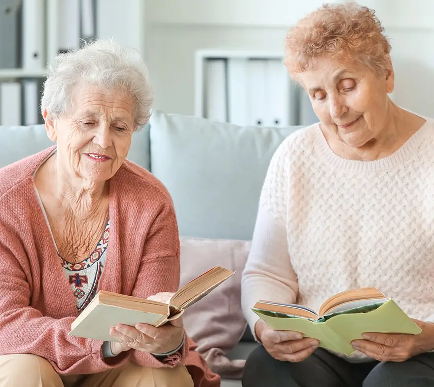 Two elderly women sitting on a couch, reading books and smiling, in a cozy living room setting.