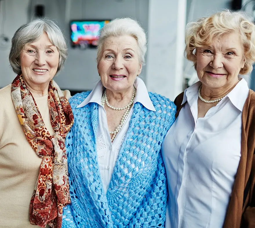 Three smiling senior women standing together, showcasing friendship and joy, in a bright indoor setting. The women are dressed in stylish outfits, with one wearing a blue cardigan and another adorned with a scarf.