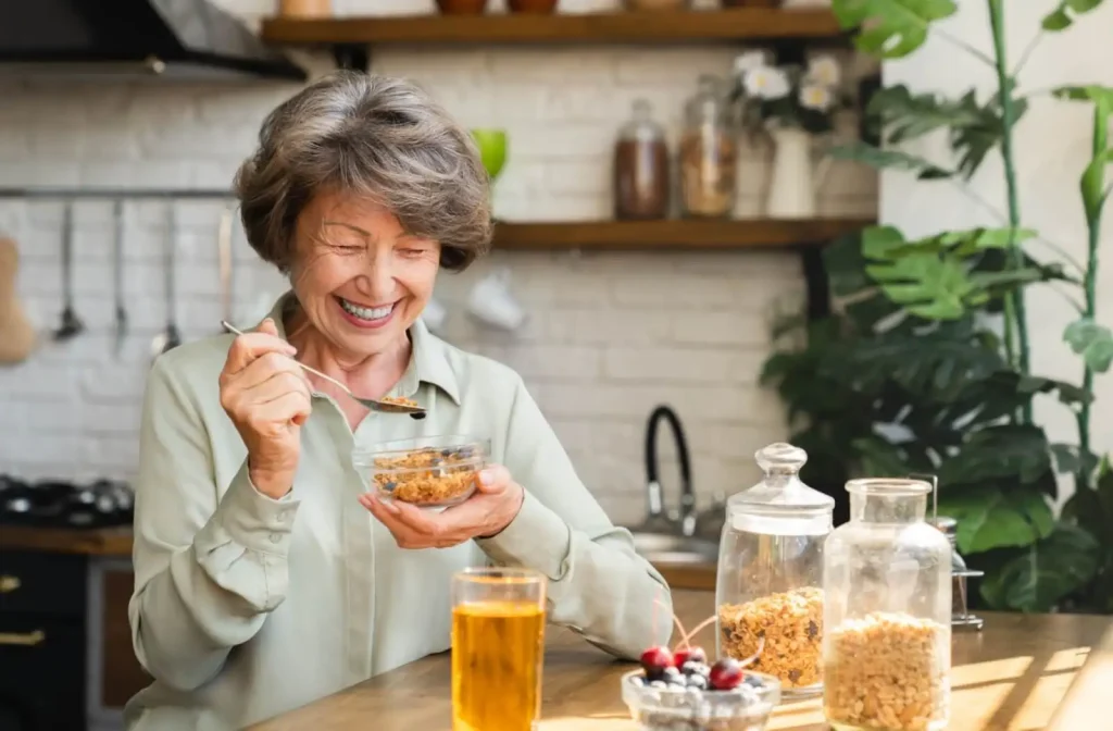Smiling elderly woman enjoying a bowl of granola at a cozy kitchen table, with a glass of iced tea and jars of ingredients in the background.