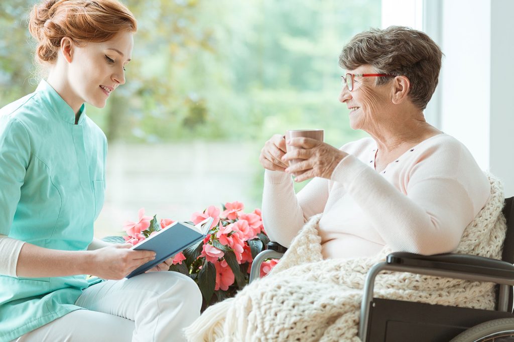 Caregiver reading to elderly woman in wheelchair, enjoying a warm drink, surrounded by flowers in a bright, inviting setting.