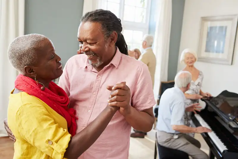Elderly couple dancing joyfully in a bright room, with a pianist and other seniors enjoying a social gathering in the background.