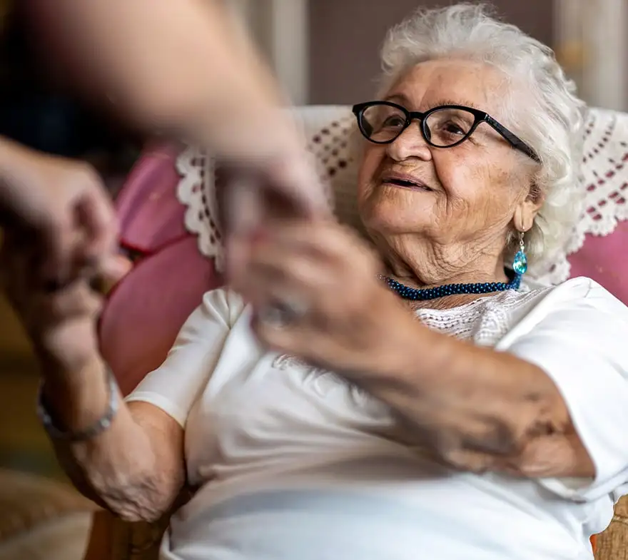 Elderly woman with white hair and glasses smiling while reaching out to hold hands with a caregiver, seated in a cozy living room setting. The scene captures a moment of warmth and connection between generations.
