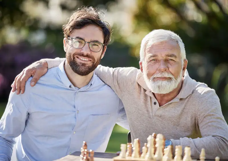 A young man and an older man smile together while sitting at a table with a chessboard in a garden setting, showcasing a moment of connection and leisure.