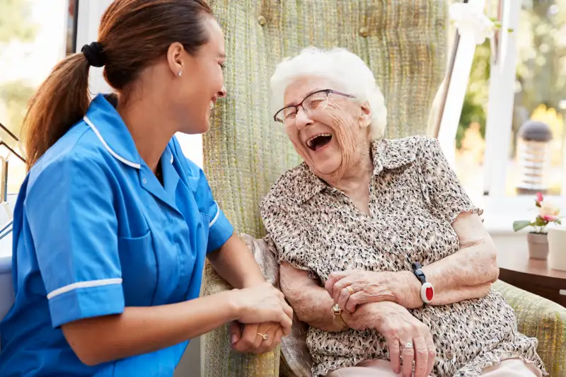 A caregiver in blue scrubs shares a joyful moment with an elderly woman in a cozy chair, both smiling and engaging in conversation, highlighting the importance of companionship and quality care in senior living.