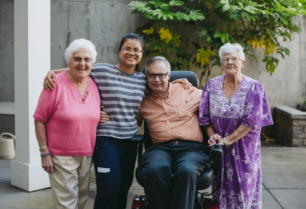 Group of four people smiling together outdoors, including two elderly women, a man in a wheelchair, and a young woman, showcasing companionship and community spirit.