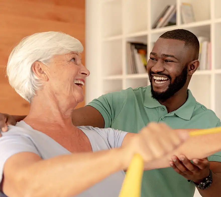 A smiling elderly woman engages in a physical therapy session with a cheerful male therapist, using a resistance band in a bright, modern living space.